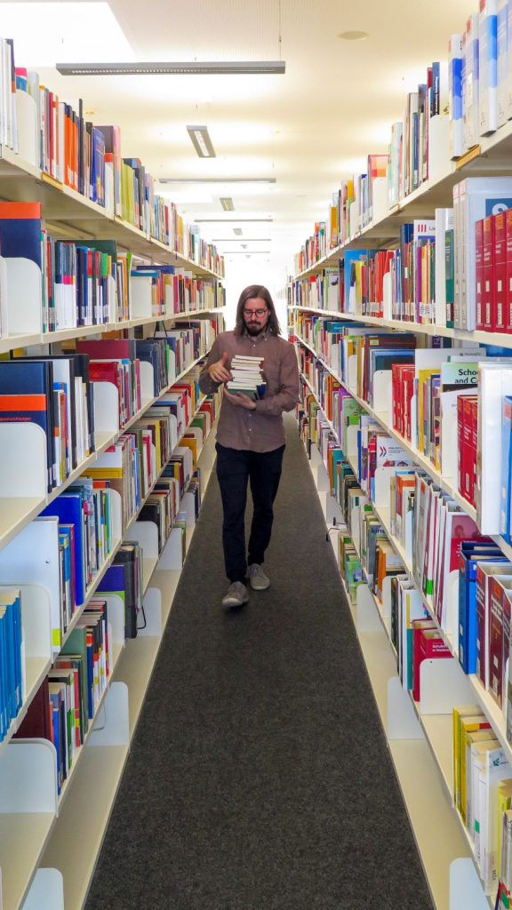 Ein Mann geht durch einen schmalen Gang zwischen zwei hohen Bücherregalen.