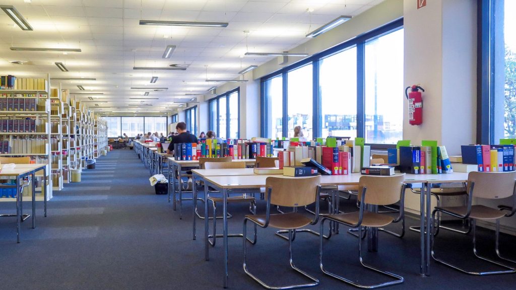 Blick in den Lesesaal Rechstwissenschaften: Links befinden sich Regalreihen mit Büchern, rechts stehen Tische mit Stühlen, an denen Studierende lernen.